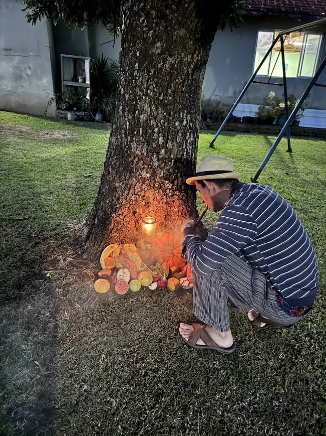 Um homem ajoelhado junto a uma árvore acende velas diante de frutas oferecidas.