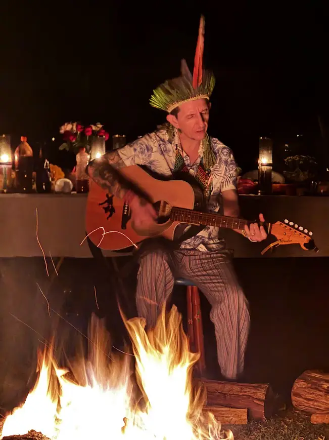 Um homem de cocar toca violão junto ao fogo, à noite.