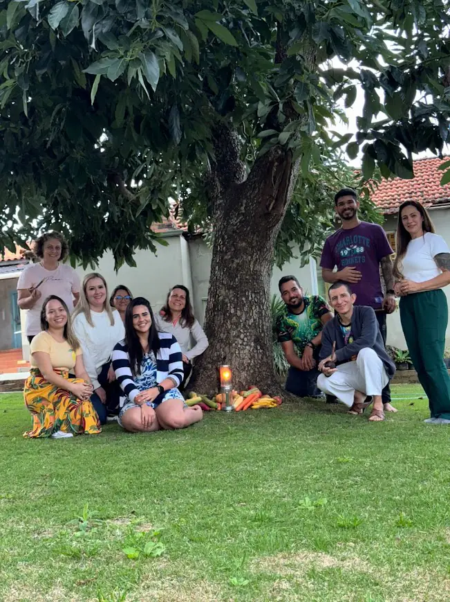 Um grupo reunido à sombra de uma árvore, sentado e em pé na grama.